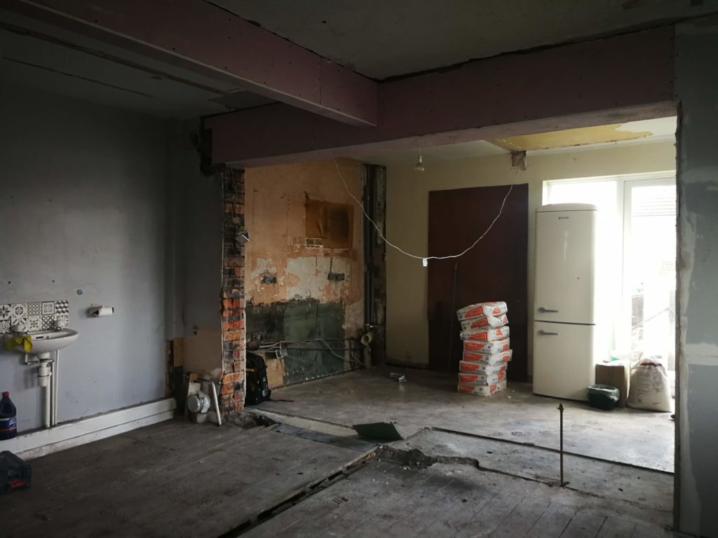 Building works in progress for an open-plan kitchen and living room remodel. Empty room with knocked down a partition wall. A basin is still visible on one wall. 
