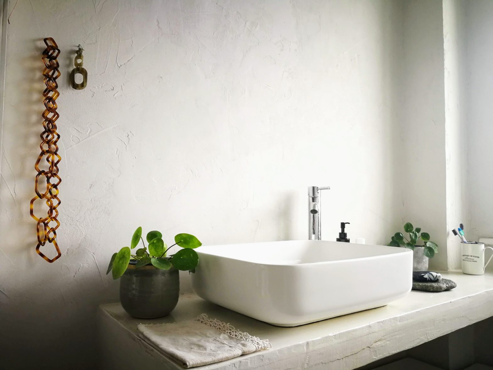 boho-chic, contemporary bathroom. Large raised basin on an L-shaped countertop. White micro-cement walls and plenty of greenery completed with a resin neckless display