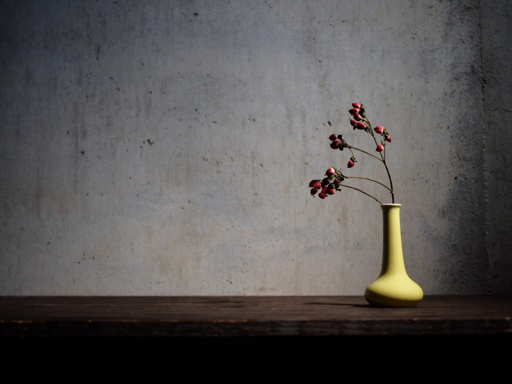 A small yellow vase with dried red berries on a wooden bench is set against a textured cement wall. The essence of WABI-SABI aesthetics 
