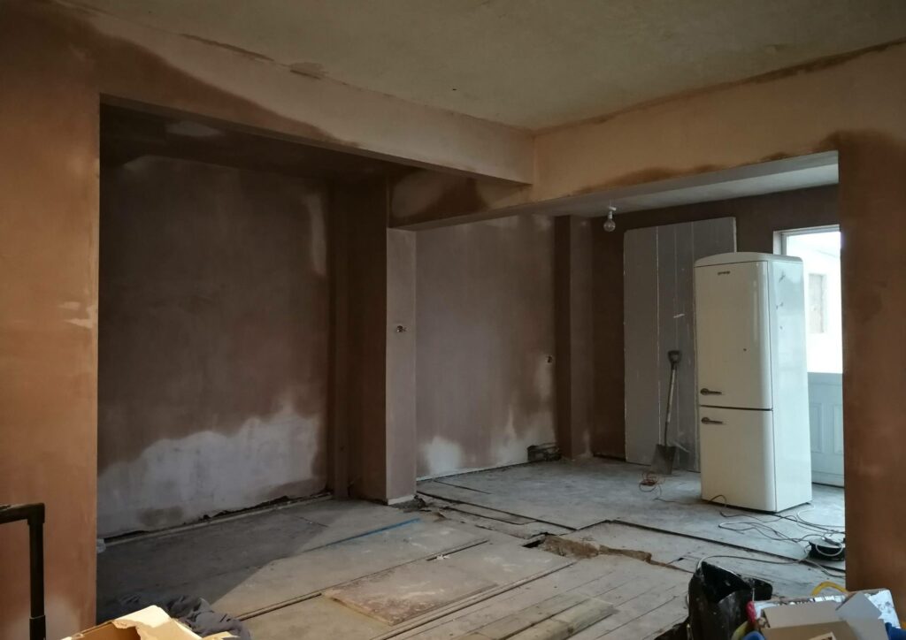 Renovation works to remodel an open plan living room. Plaster is drying on the walls and a tall white fridge is in the middle of the room