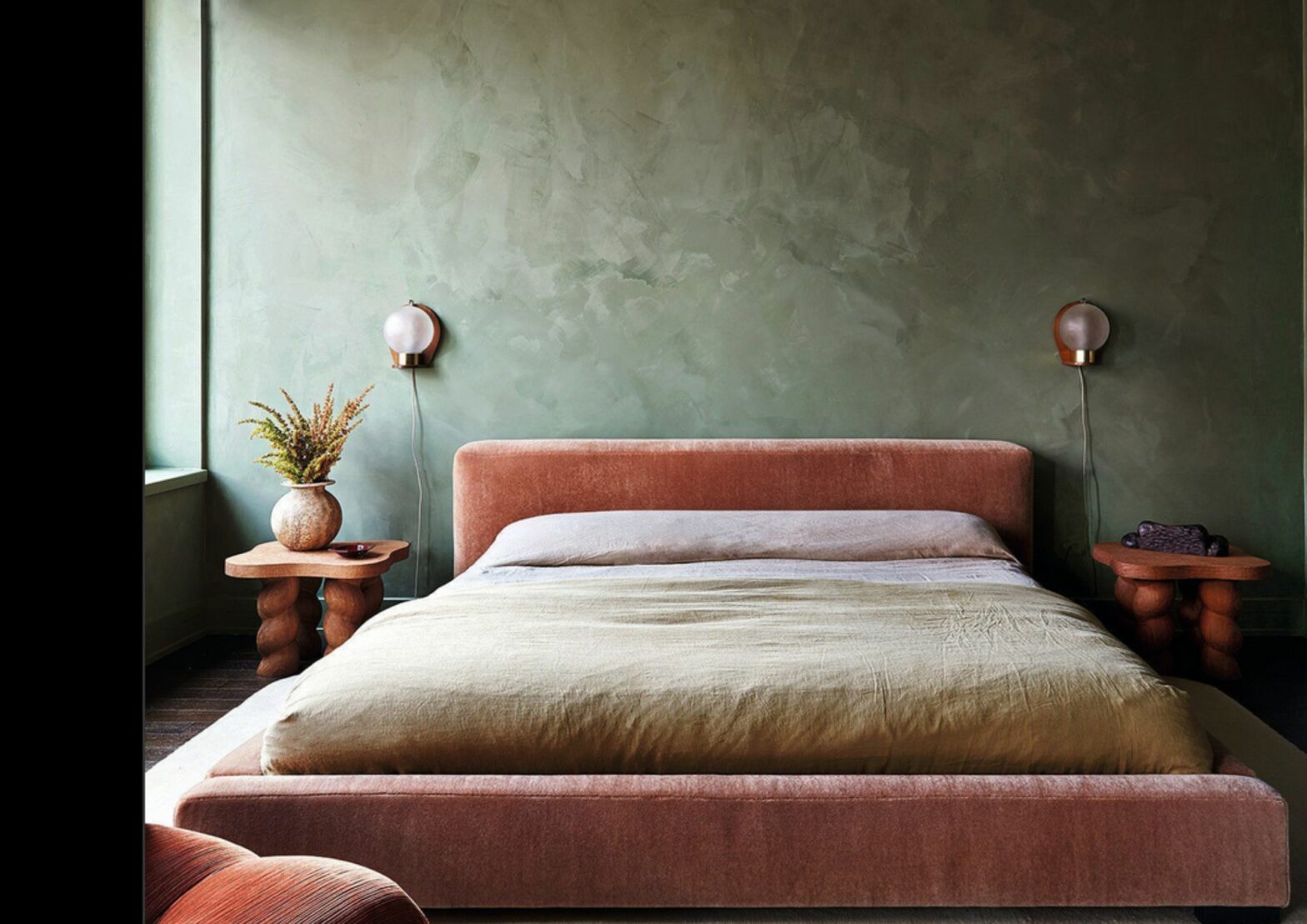 Contemporary bedroom with green limewash walls and soft pink upholstered bed. Two wall-mounted bedside bulbs and a vintage wooden bedside table.